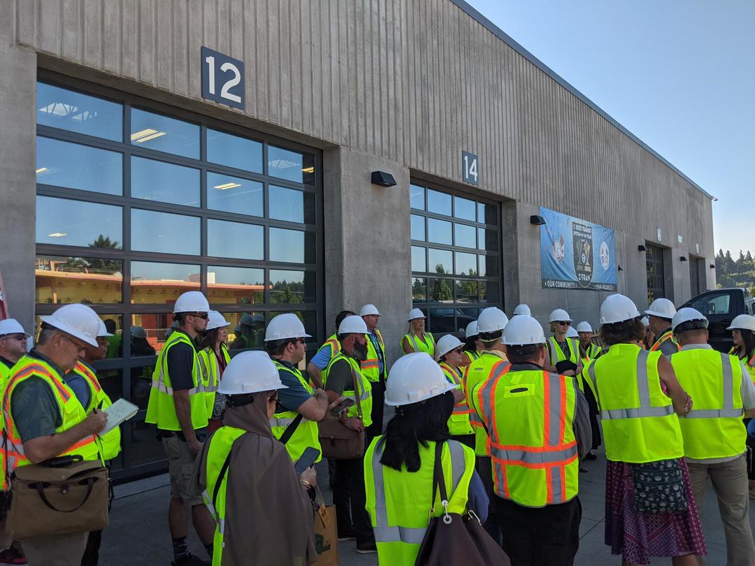 a group of transit professionals in hard hats and safety vests gather outside a maintenance building